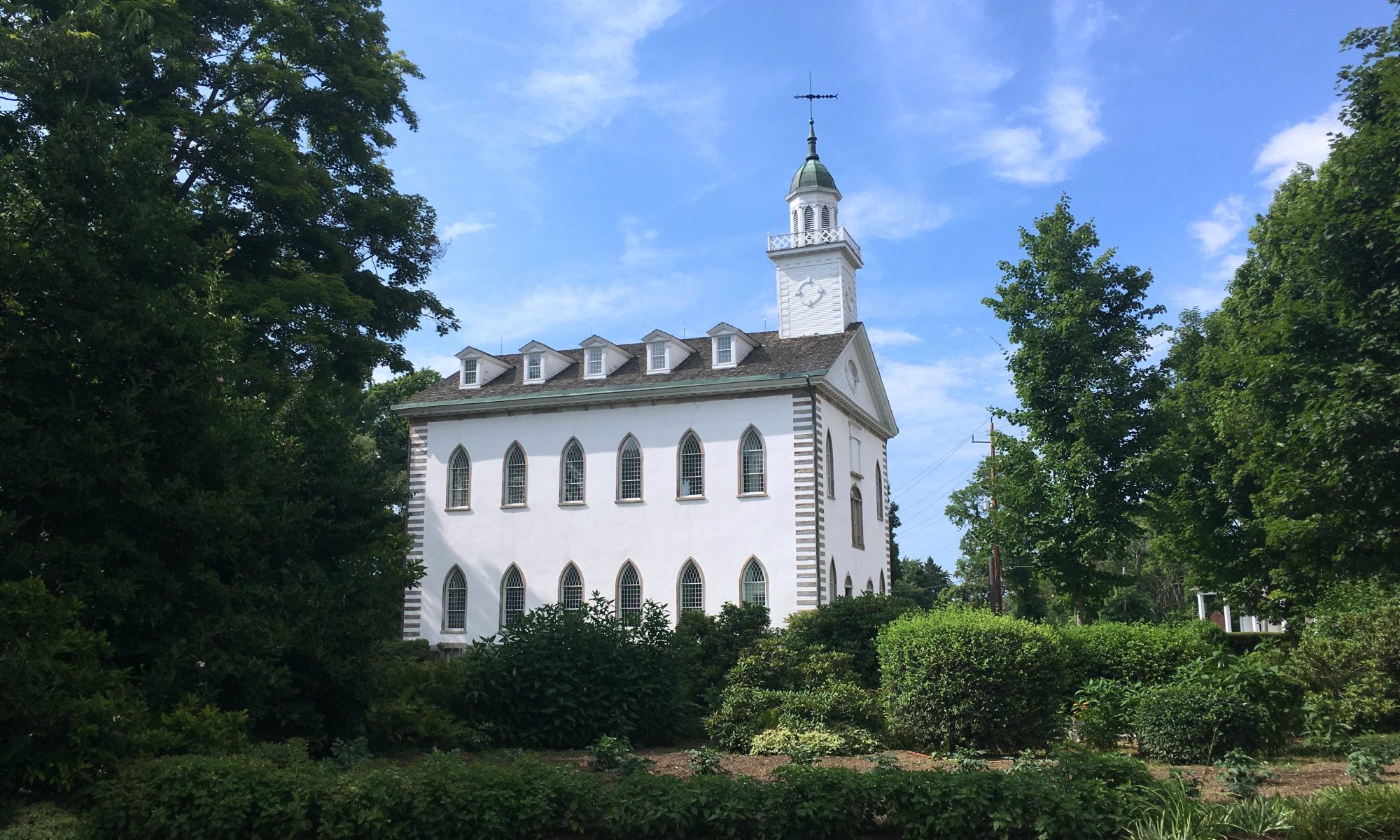 The Kirtland Temple, a white building with two stories and a garret and bell tower, is seen from across a lawn, bushes and trees.