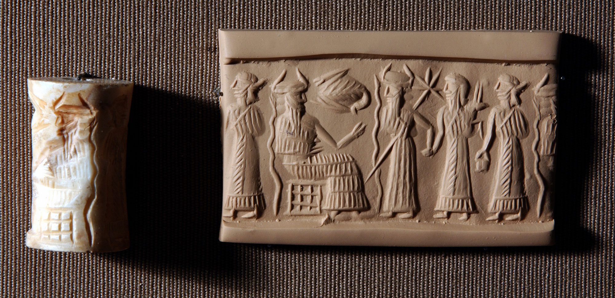 A white seal in the shape of a cylinder with sculpted indentations around its circumference and a block of clay on which it was used sit on a surface. The image on the block of clay demonstrates how the cylinder seal works. The image appears to be of several people standing in front of one figure sitting on a throne.