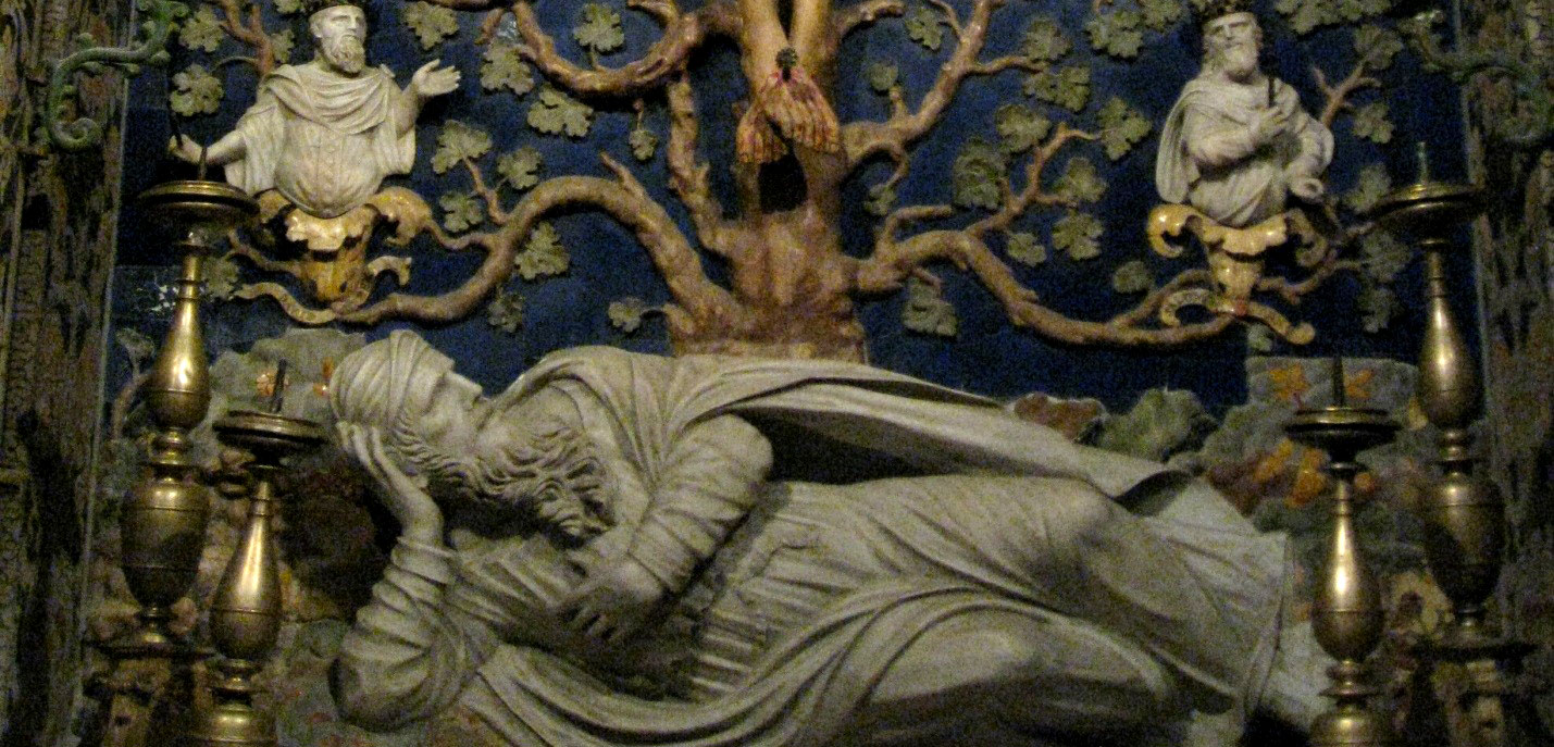A sculpture of a man in clothing and a cloak, perhaps in marble, lies reclined in front of a sculpture of a gnarled tree. Two of the branches of the tree hold up at their ends a blossom, inside each of which is the sculpture of the clothed upper body of a man.