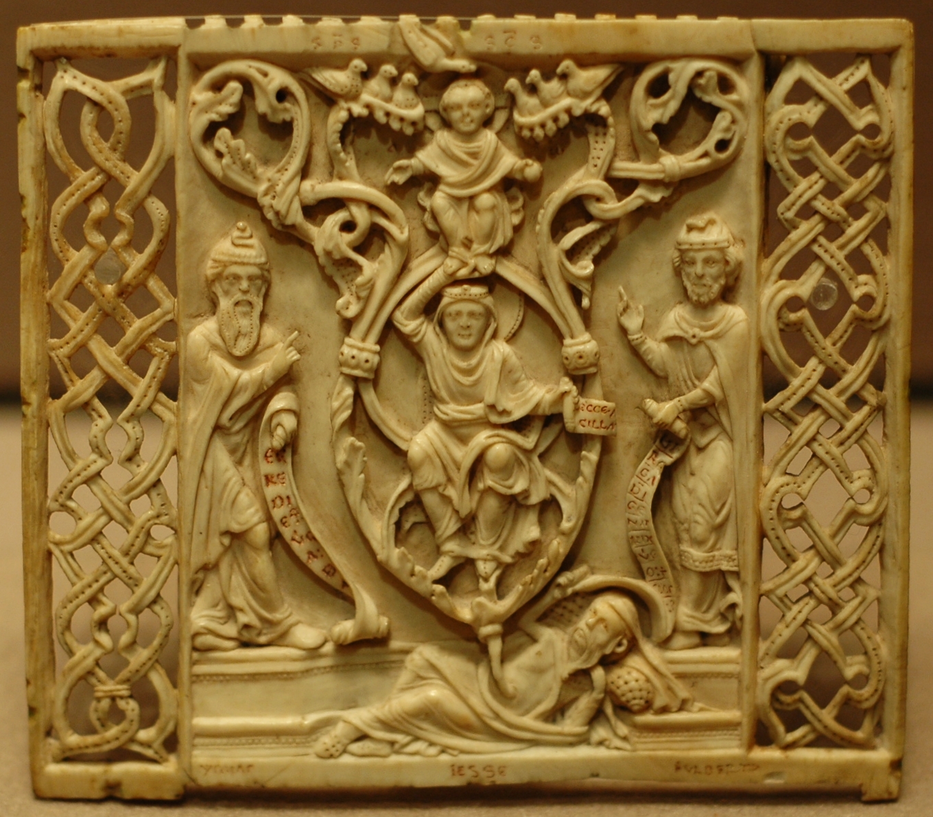 An ivory sculpture. a man lying on the ground appears to have a root protruding from his bellybutton and stretching into branches. In the branches are a man holding a scroll, a child, and several birds. The branches are flanked on either side by men holding scrolls. The sculpture is very ornate.