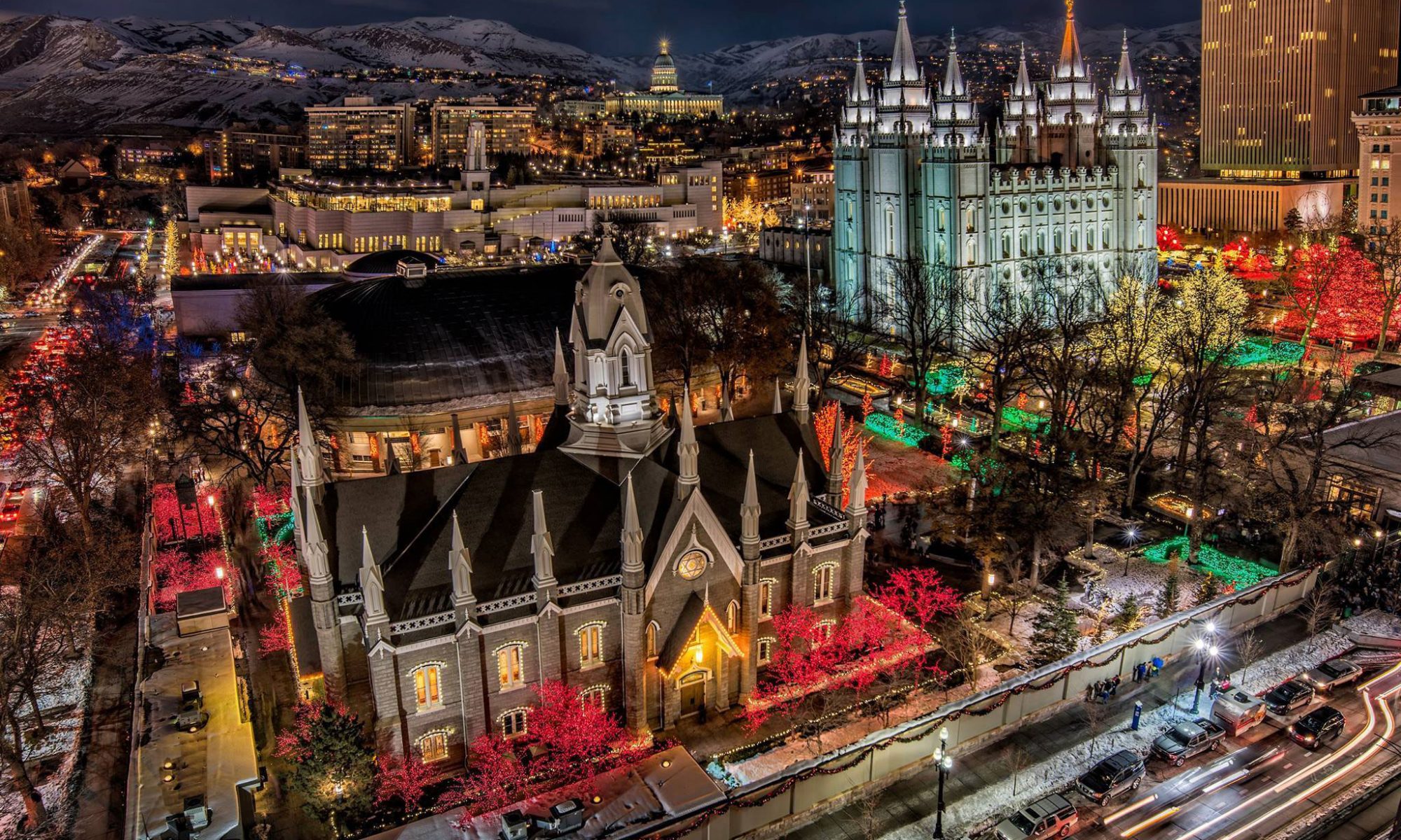 An image of Temple Square at Christmas. In the foreground is the chapel on temple square, and in the background is the temple. All around in the trees and bushes are bright Christmas lights. Other Salt Lake City building are visible in the background behind the temple.