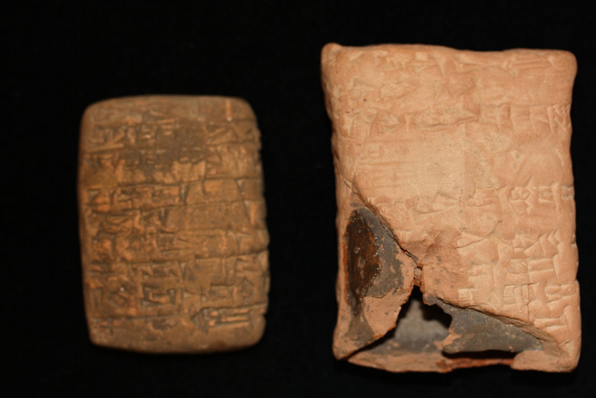 A dark clay tablet sits on a surface next to a lighter clay container. Both are etched with cuneiform characters.