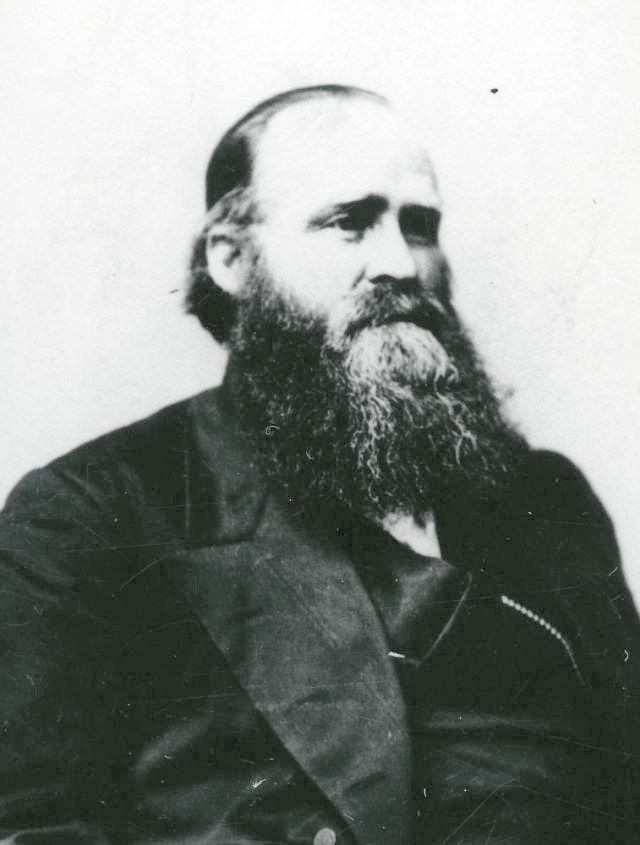 A photograph of Amasa Lyman. He wears a longer beard and mustache, is balding, and seems to have a large frame. He wears a dark coat and vest and white shirt.
