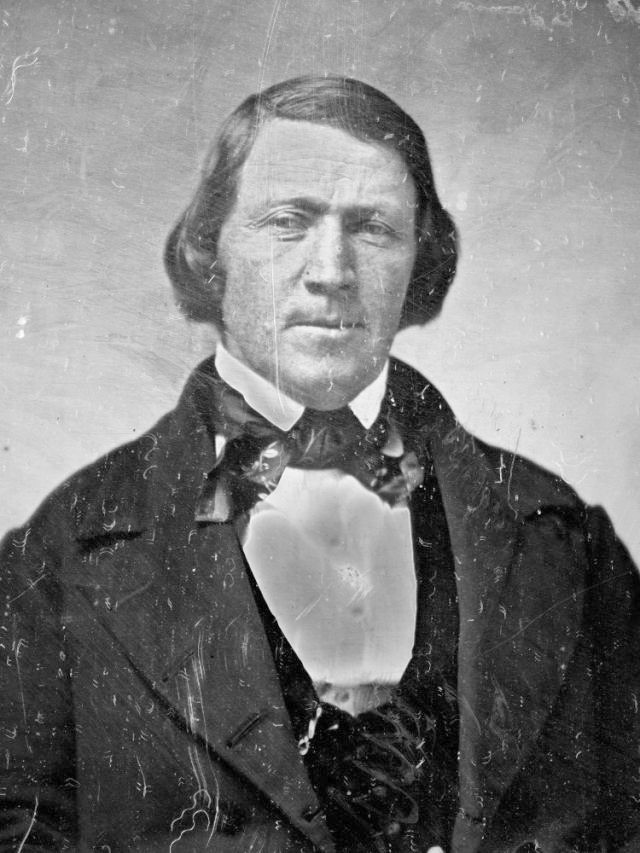 A daguerrotype of Brigham Young. He wears a dark coat, a darker vest, a white shirt, and a dark necktie or cravat. His hair is parted to one side and sort of curls at his cheeks.