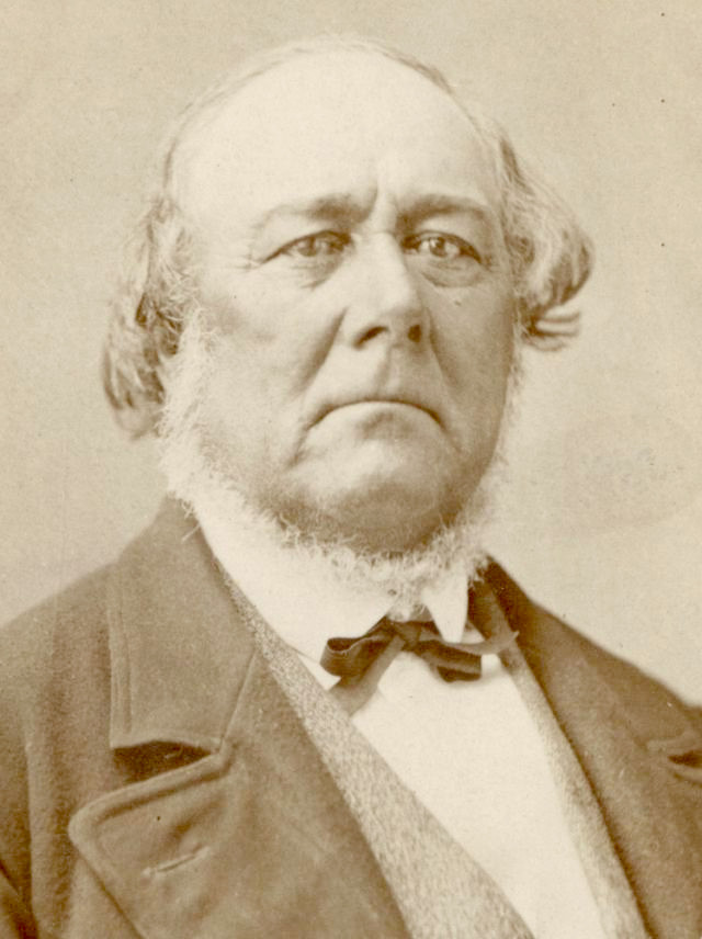 A sepia photo of Charles C. Rich. He wears a dark coat, light vest, white shirt, and dark tie. He has a light-colored neck beard. He has wavy, light hair but is balding.