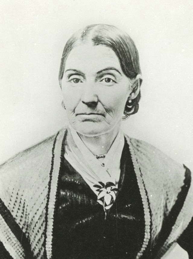A photograph of Emma Smith. She wears a thin white scarf with a flower brooch underneath a dark blouse and a light-colored, patterned shawl. Her hair appears to be brought back into a bun. She has age lines around her mouth and wears a neutral expression.