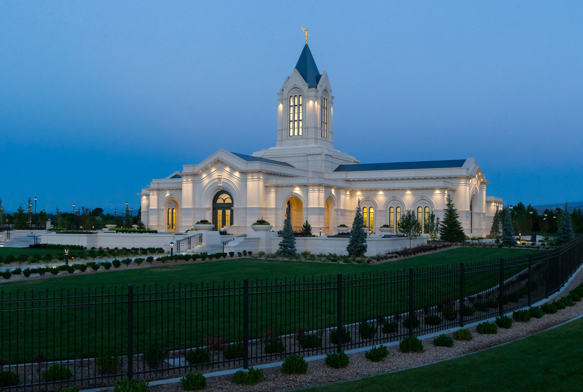 An image of the Fort Collins Colorado Temple. The temple is online one floor tall but has a tower in the middle. The photograph was taken in the evening, and the sky and lawn are darkening, while all the lights in the temple are on.