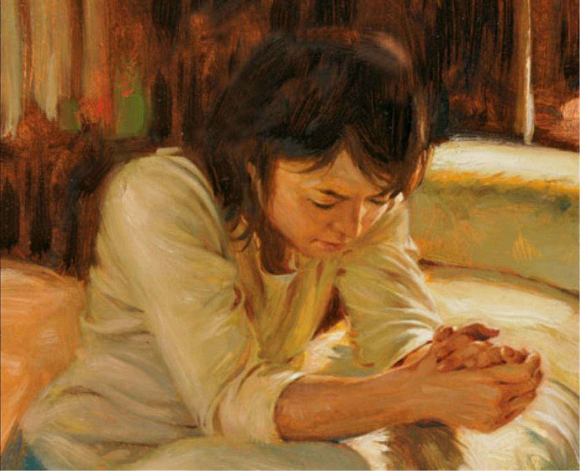 A woman kneels next to a bed. Her hands are clasped together, and her arms are on the bed as she bows her head in prayer. She wears a white shirt and has dark hair.